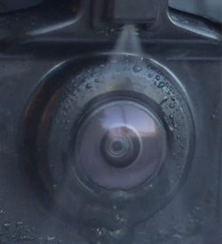 Photograph of standard fixed nozzle spraying fluid to clear camera lens on vehicle.